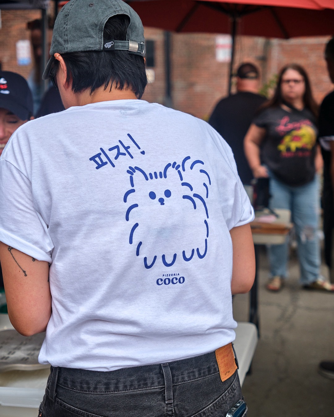 Back of a Pizzeria Coco business T-shirt featuring a cute animated Coco the dog at an event.