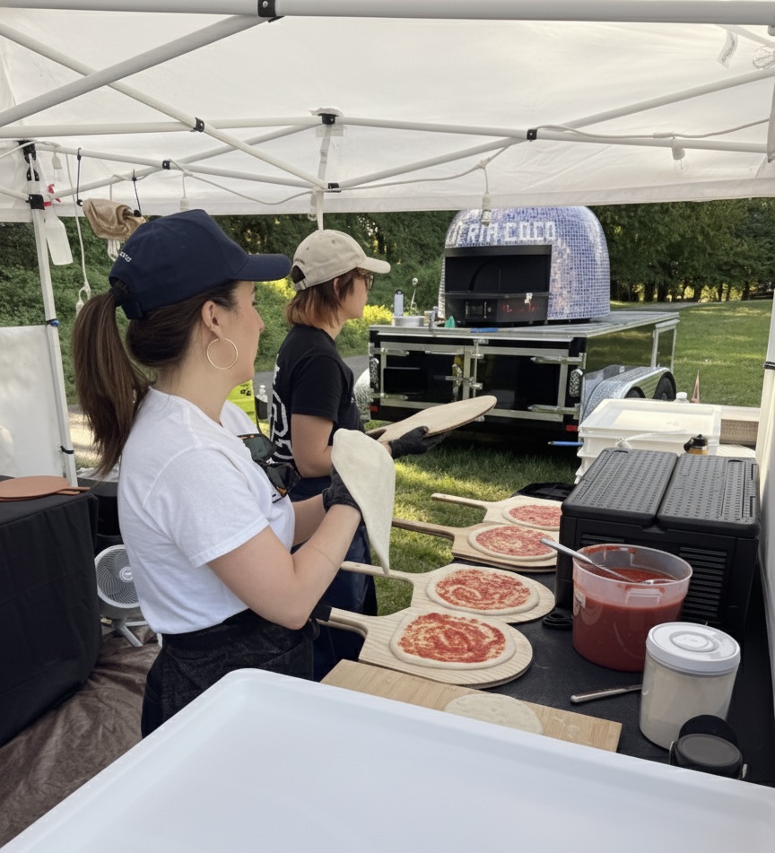 Chefs making fresh pizzas onsite at a daytime outdoor event, with dough stretched and ingredients ready on the prep table at a Pizzeria Coco catering event.